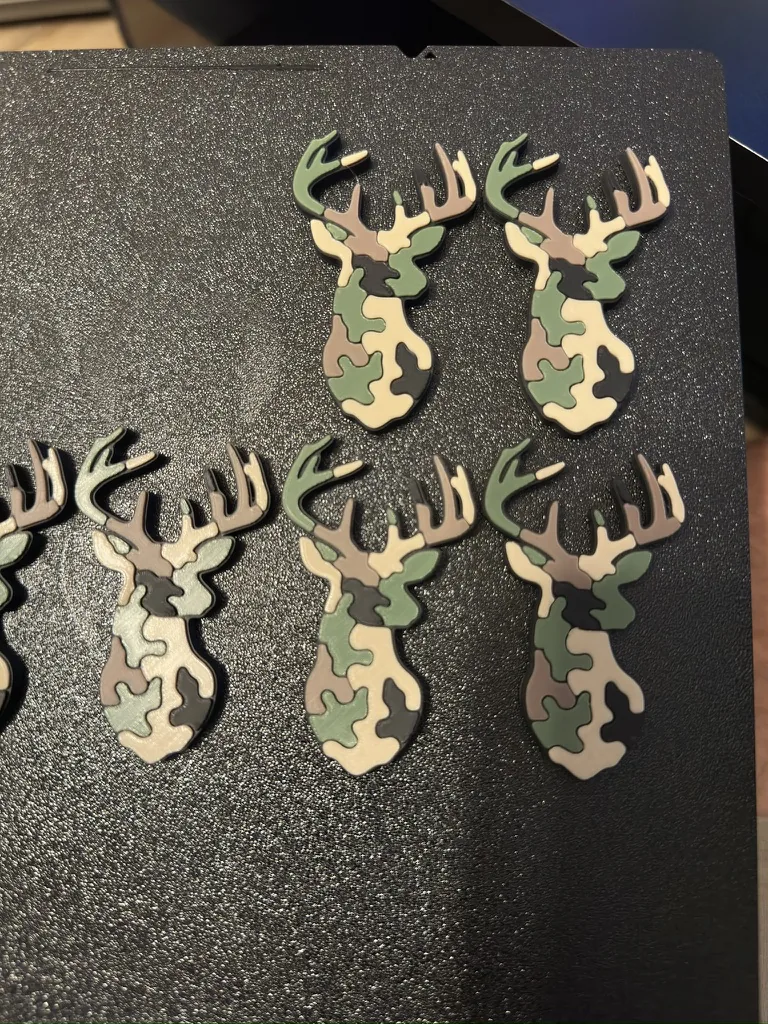 Cinq porte-clés en forme de tête de cerf, finition camouflage vert et marron, alignés sur le plateau d'une imprimante 3D