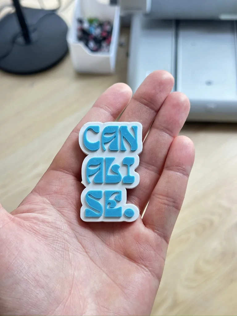 Porte-clé bleu marine au logo CANALISE. en relief blanc, tenu en main devant l'imprimante 3D de l'atelier Filon Médoc