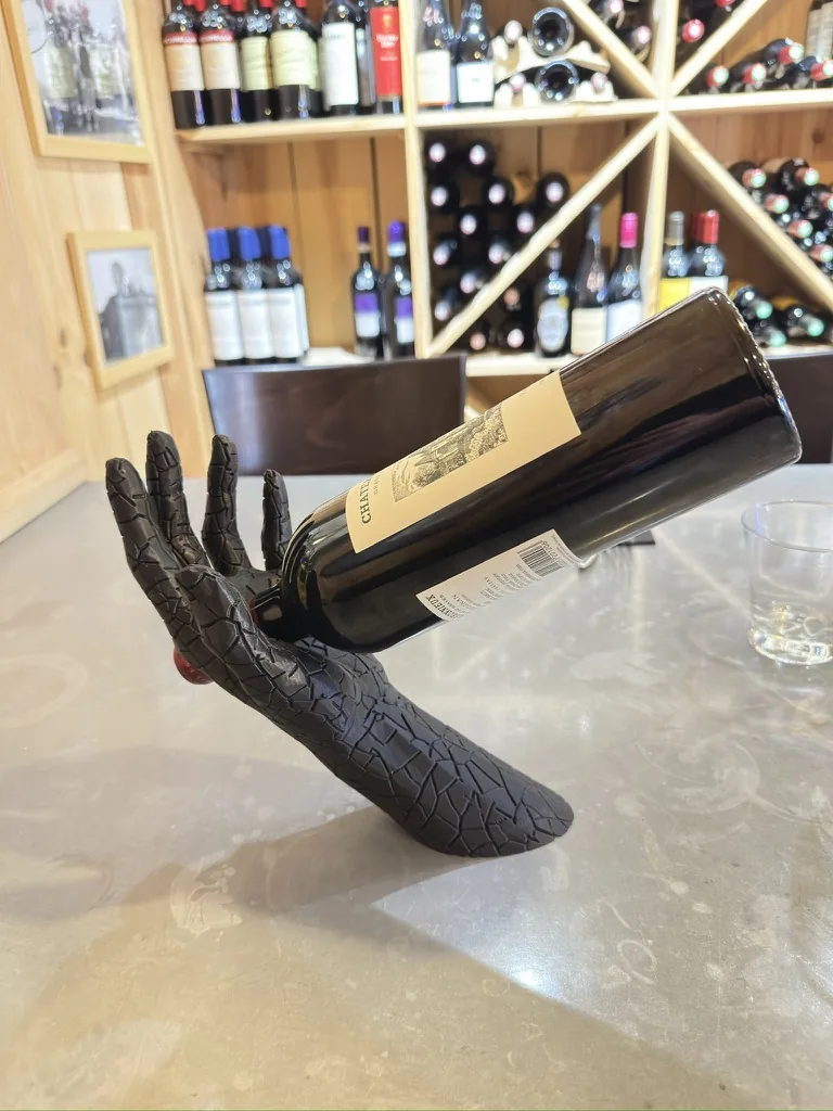 Support de bouteille noir mat en forme de main de gargouille tenant une bouteille de vin, posé sur un meuble en bois sombre